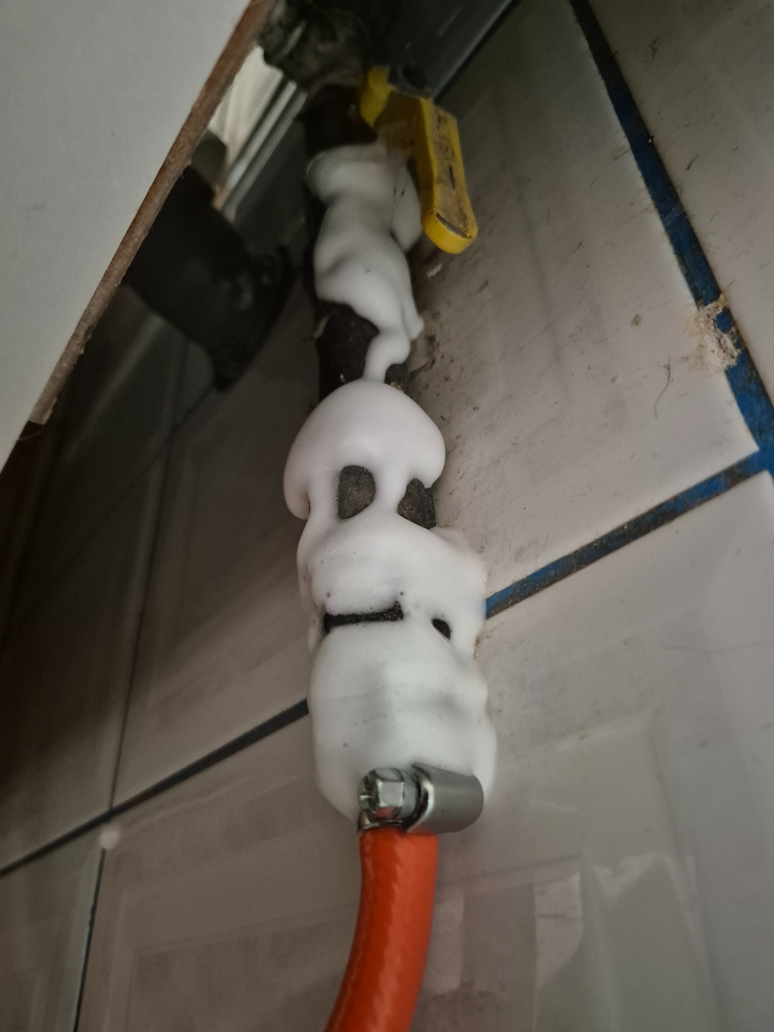 Czarna rura gazowa z żółtym zaworem, pokryta pianą do wykrywania nieszczelności, z podłączonym pomarańczowym wężem, na tle białych płytek z niebieską fugą.