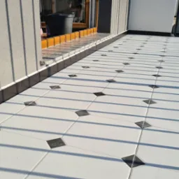 Wyłożony białymi płytkami z czarnymi akcentami balkon, z widocznymi cieniami barierki i fragmentem ściany budynku.