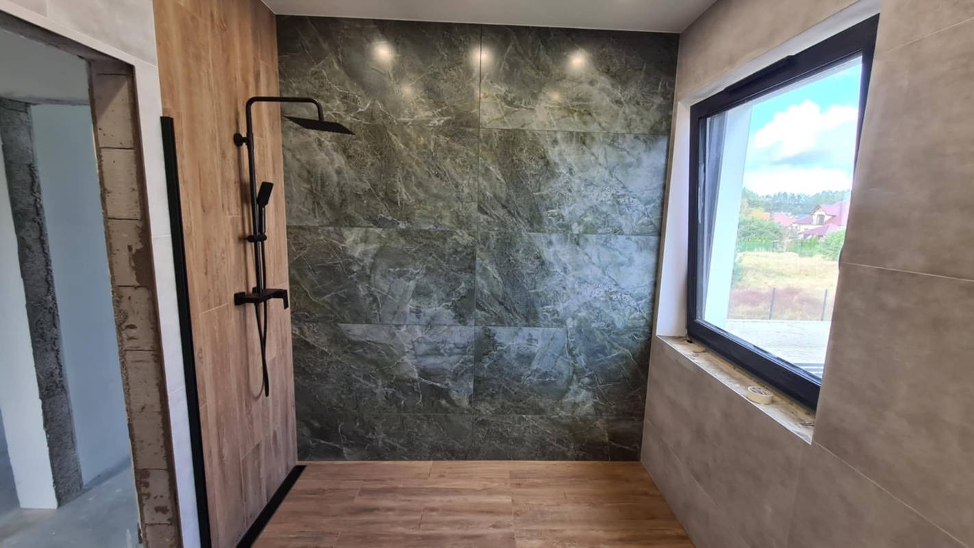 Nowoczesna kabina prysznicowa z czarną armaturą, wyłożona płytkami imitującymi drewno i marmur, z oknem w czarnej ramie.
