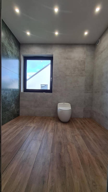 Nowoczesna łazienka w Gdańsku z drewnopodobną podłogą, szarą płytką na ścianach i wiszącą toaletą, widziana z poziomu podłogi, z oknem w tle.