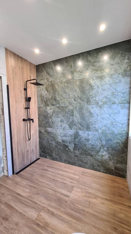 Nowoczesna kabina prysznicowa walk-in z czarną armaturą i płytkami imitującymi drewno na ścianie oraz podłodze, zielone płytki marmurowe na ścianie prysznicowej, oświetlenie punktowe w suficie.