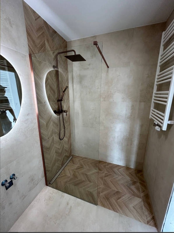 Nowoczesna łazienka z kabiną prysznicową typu walk-in, wykończona beżowymi płytkami w dwóch odcieniach i wzorach, z deszczownicą w kolorze miedzi oraz okrągłym lustrem z podświetleniem LED.