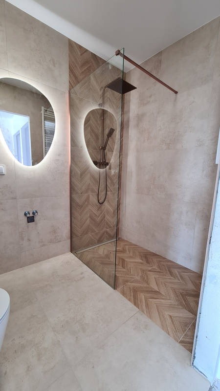 Nowoczesna łazienka z kabiną prysznicową typu walk-in, wykończona beżowymi płytkami i drewnopodobną mozaiką w jodełkę na podłodze i ścianie kabiny. Okrągłe lustro z podświetleniem LED zawieszone...