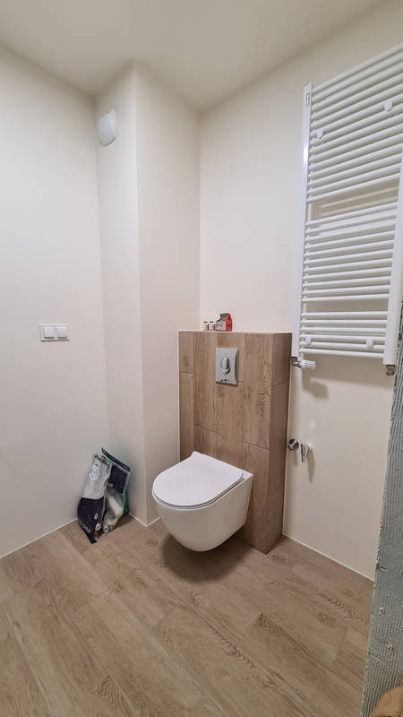 Nowoczesna toaleta podwieszana z zabudową z płytek imitujących drewno, biały grzejnik drabinkowy na ścianie, podłoga z paneli w odcieniach beżu, worki z materiałami budowlanymi w rogu.