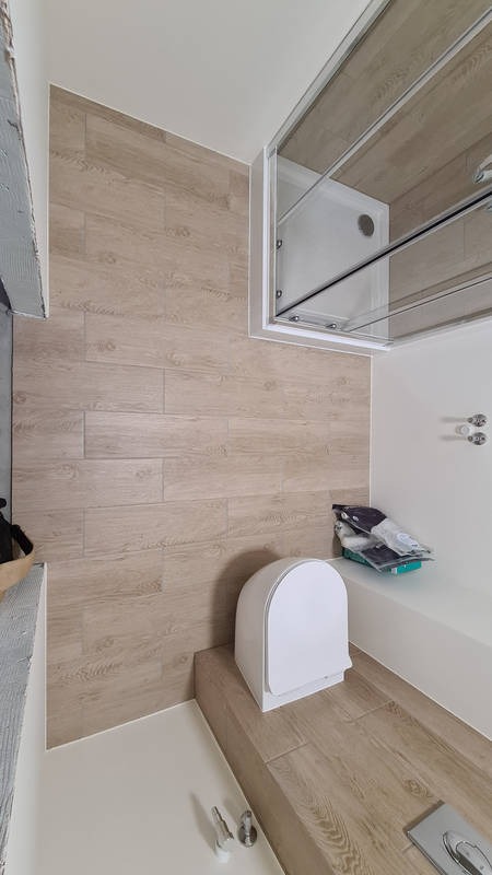 Nowoczesna łazienka po remoncie z drewnopodobnymi płytkami na ścianach i podłodze, kabina prysznicowa z przesuwnymi drzwiami, wisząca toaleta z deską sedesową typu slim, widoczne akcesoria...