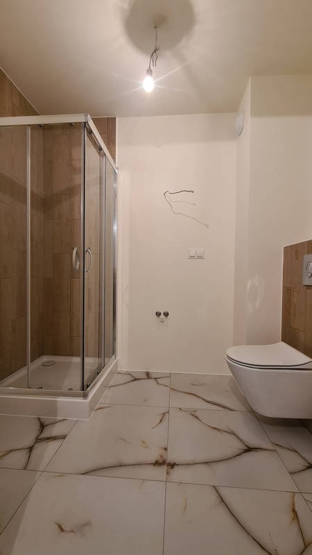 Niedokończona łazienka z kabiną prysznicową, białymi płytkami imitującymi marmur z brązowymi żyłami, wiszącą toaletą i wystającymi kablami ze ściany.