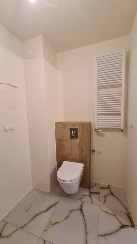 Nowoczesna toaleta podwieszana z drewnopodobną zabudową stelaża i białym grzejnikiem drabinkowym na beżowej ścianie, marmurowe płytki podłogowe z brązowymi żyłami.