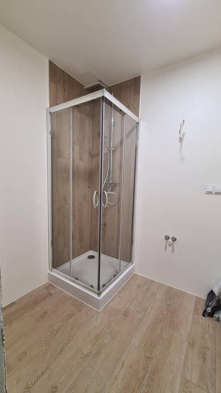 Nowoczesna kabina prysznicowa z przesuwnymi drzwiami w narożniku łazienki, wykończona płytkami imitującymi drewno, z widoczną armaturą i odpływem liniowym, na podłodze panele podłogowe w odcieniach...