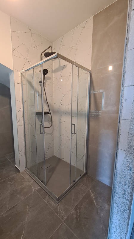 Kabina prysznicowa narożna z chromowanymi profilami, wykończona płytkami imitującymi marmur na ścianach i podłodze, widoczny fragment nieotynkowanej ściany z prawej strony.
