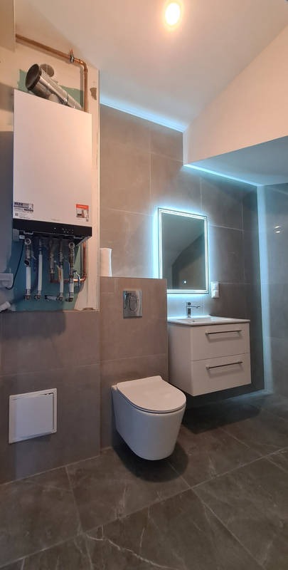 Nowoczesna łazienka z szarymi płytkami imitującymi marmur, podwieszana toaleta, biała szafka z umywalką i podświetlanym lustrem, wiszący piec gazowy z widocznymi rurami.