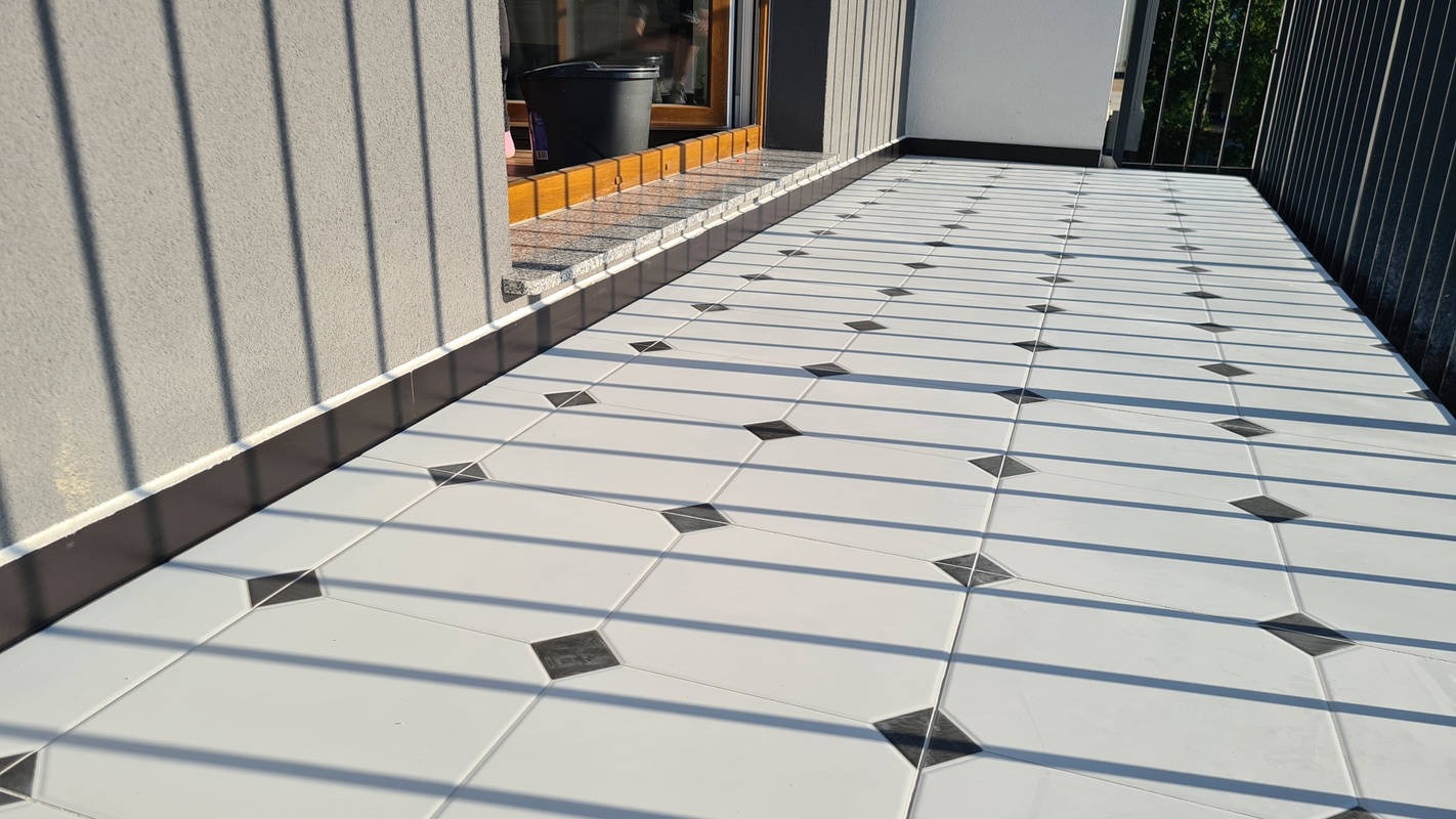 Wyłożony białymi płytkami z czarnymi akcentami balkon, z widocznymi cieniami barierki i fragmentem ściany budynku.