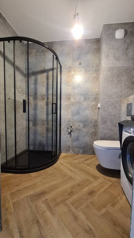 Nowoczesna łazienka z kabiną prysznicową z czarnymi profilami, płytkami imitującymi kamień na ścianach, drewnopodobną podłogą ułożoną w jodełkę i wiszącą toaletą.