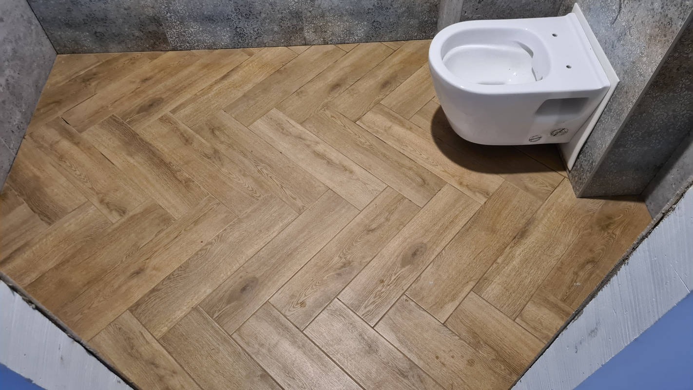 Ułożona w jodełkę podłoga z płytek imitujących drewno w łazience, z wiszącą muszlą klozetową i fragmentem szarej ściany z dekoracyjnymi płytkami.