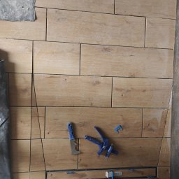 Technika Budowlana Konrad Kołecki - Układanie płytek imitujących drewno na ścianie, z widocznym odpływem liniowym i narzędziami glazurniczymi, w trakcie montażu.