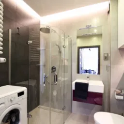 Nowoczesna łazienka z kabiną prysznicową z zaokrąglonym szkłem, pralką z przodu i podświetleniem LED na suficie, widok na wnętrze przez lustro.