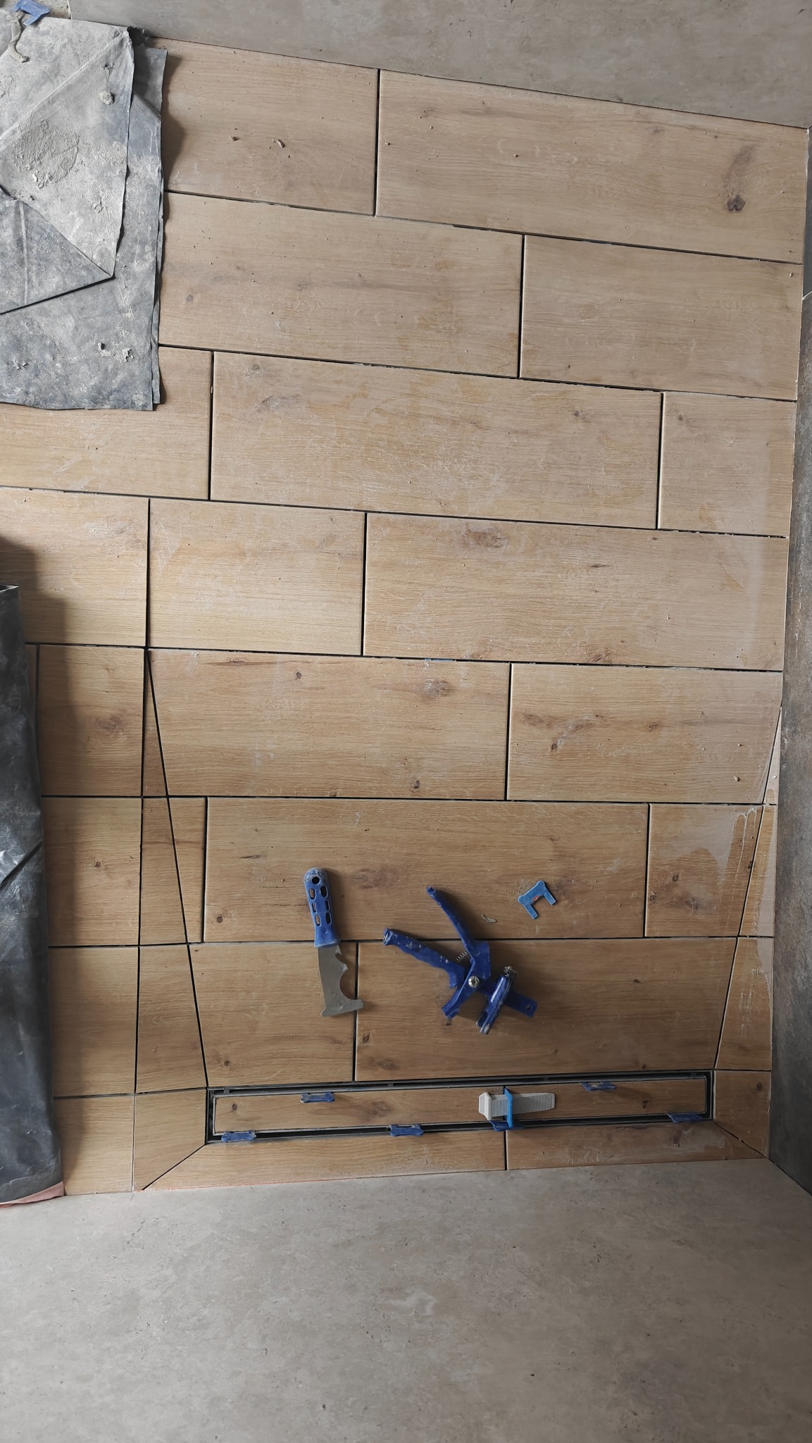 Układanie płytek imitujących drewno na ścianie, z widocznym odpływem liniowym i narzędziami glazurniczymi, w trakcie montażu.