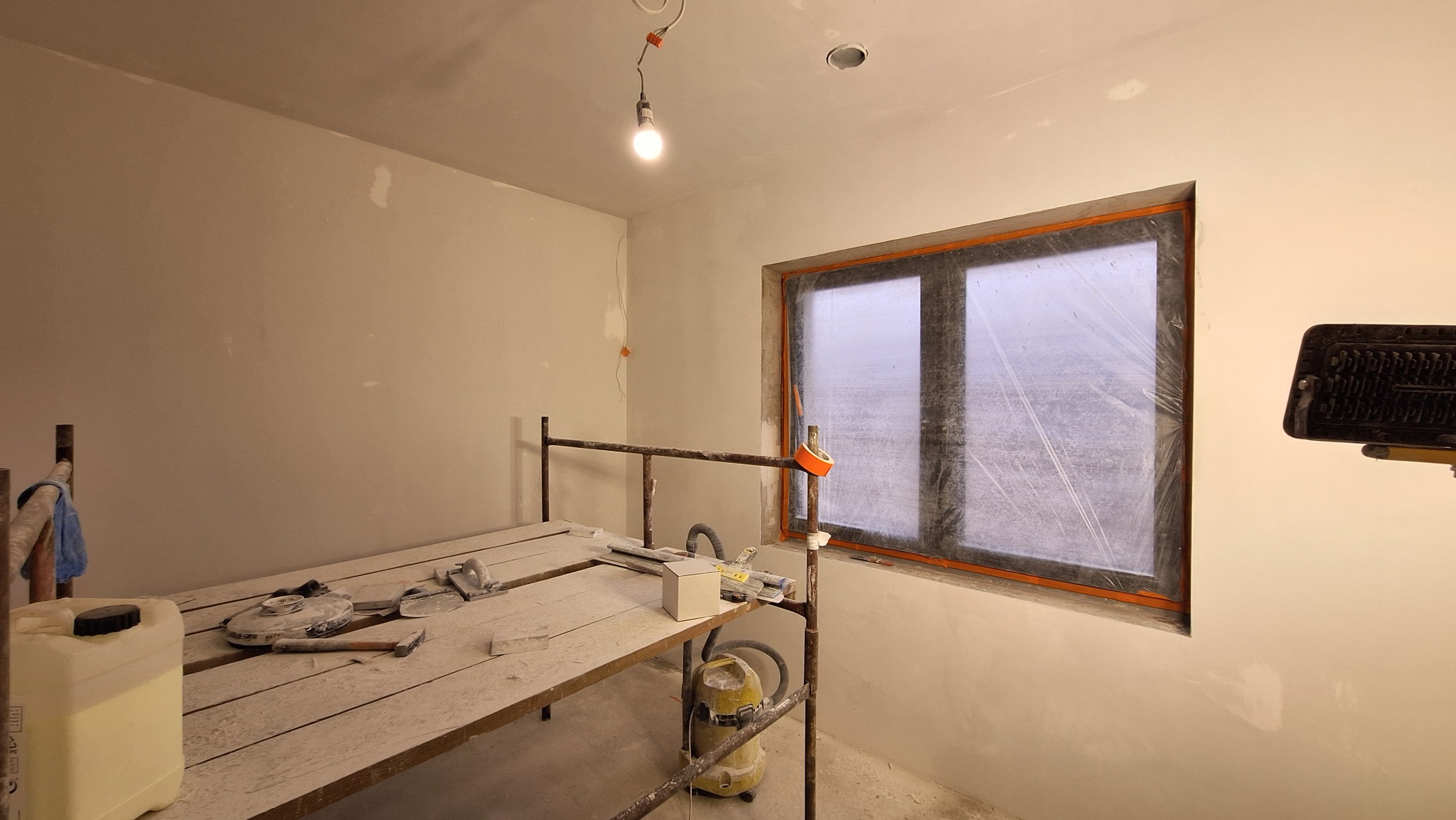 Wnętrze pokoju w trakcie prac wykończeniowych, ściany przygotowane pod malowanie, okno zabezpieczone folią, widoczne rusztowanie z narzędziami i odkurzacz przemysłowy.