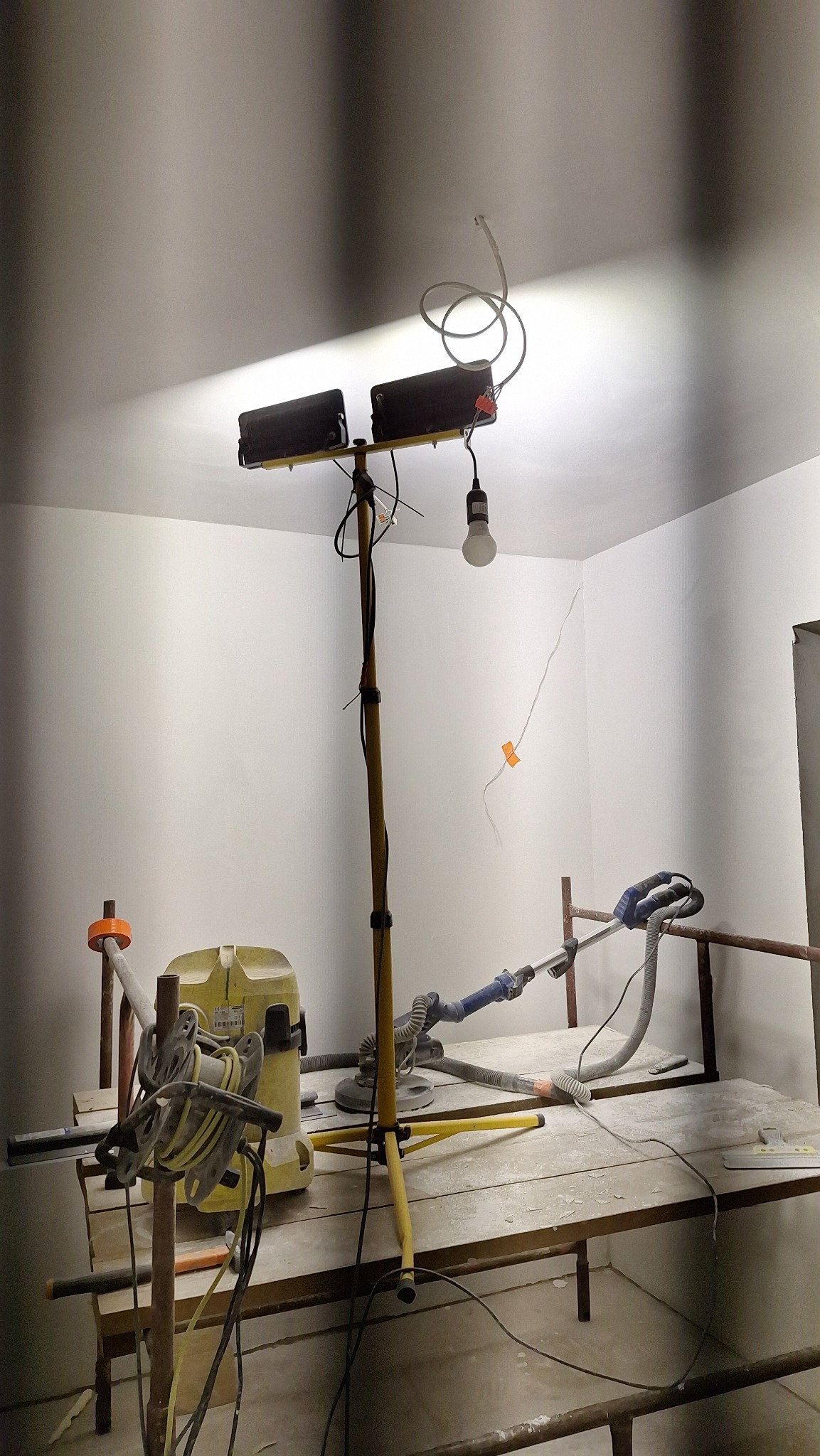 Przygotowanie do szlifowania ściany: oświetlenie halogenowe na statywie, szlifierka z odkurzaczem na rusztowaniu, widoczna taśma maskująca na pęknięciu ściany.