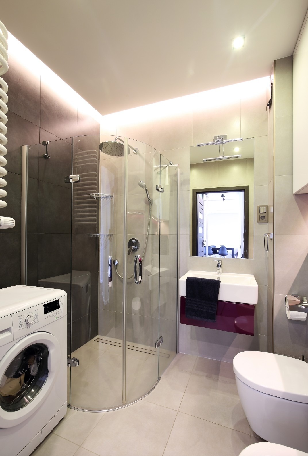 Nowoczesna łazienka z kabiną prysznicową z zaokrąglonym szkłem, pralką z przodu i podświetleniem LED na suficie, widok na wnętrze przez lustro.