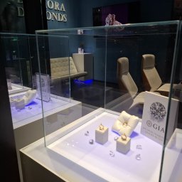 Diamora Diamonds - Ekskluzywny salon jubilerski z złotymi pierścionkami i diamentami w szklanych gablotach, oświetlonych subtelnym, niebieskawym światłem, z minimalistycznym wystrojem i logo GIA.