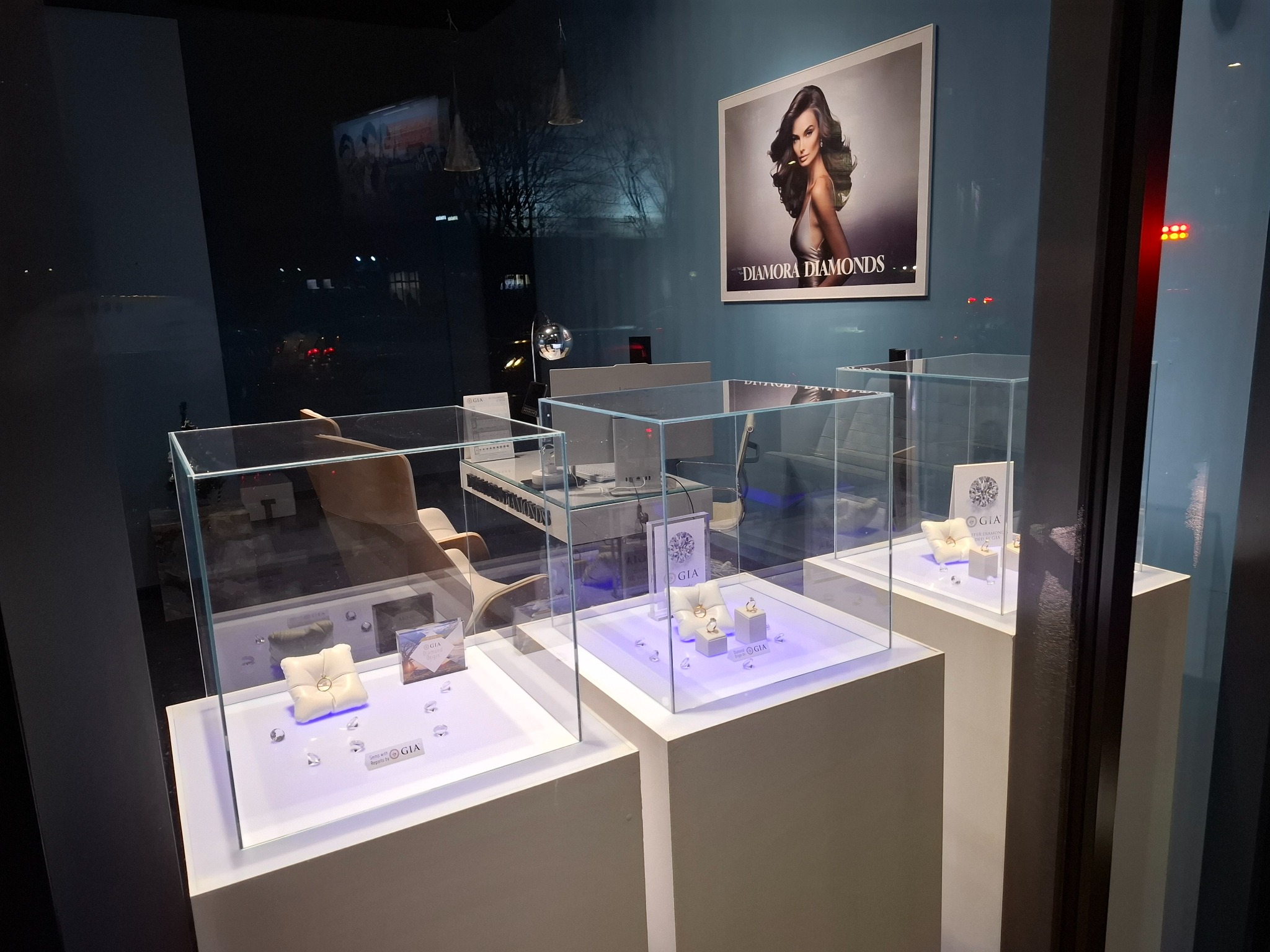 Wystawa jubilerska za szybą, prezentująca pierścionki z brylantami na białych poduszkach i platformach podświetlonych na niebiesko, z logo GIA widocznym na ekspozytorach i plakat z modelką w tle.