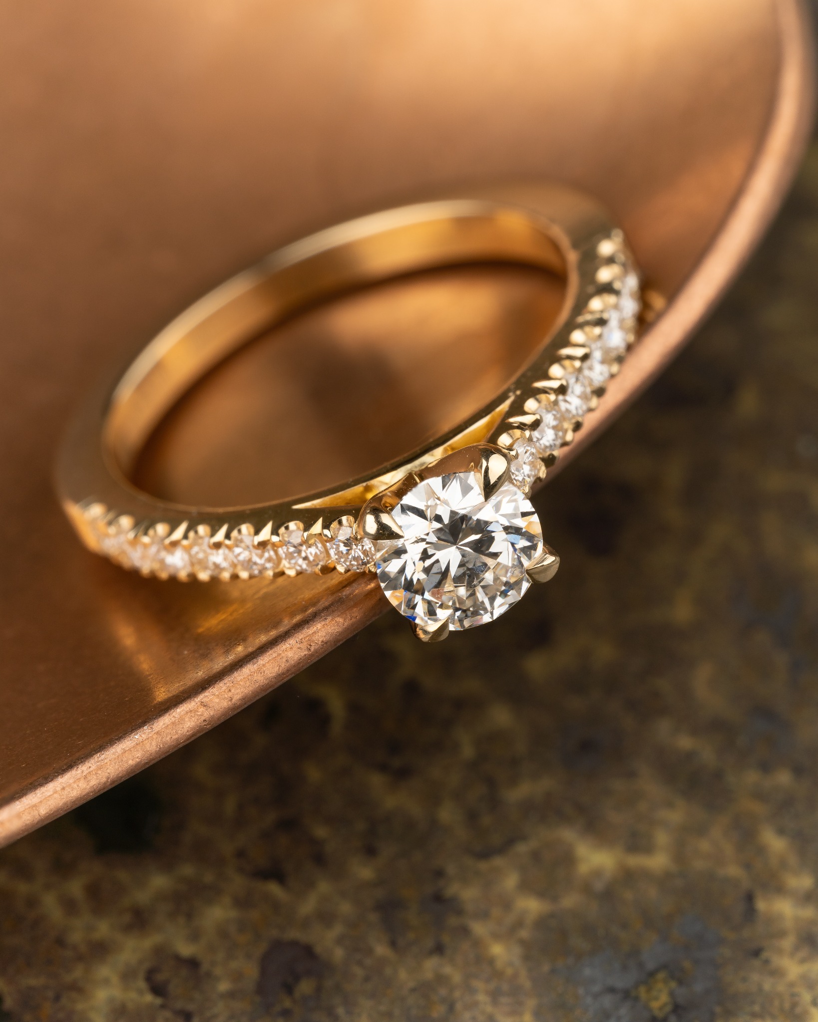 Złoty pierścionek zaręczynowy z brylantem, dodatkowo zdobiony mniejszymi kamieniami na obrączce, leżący na miedzianej powierzchni o nieregularnej fakturze.