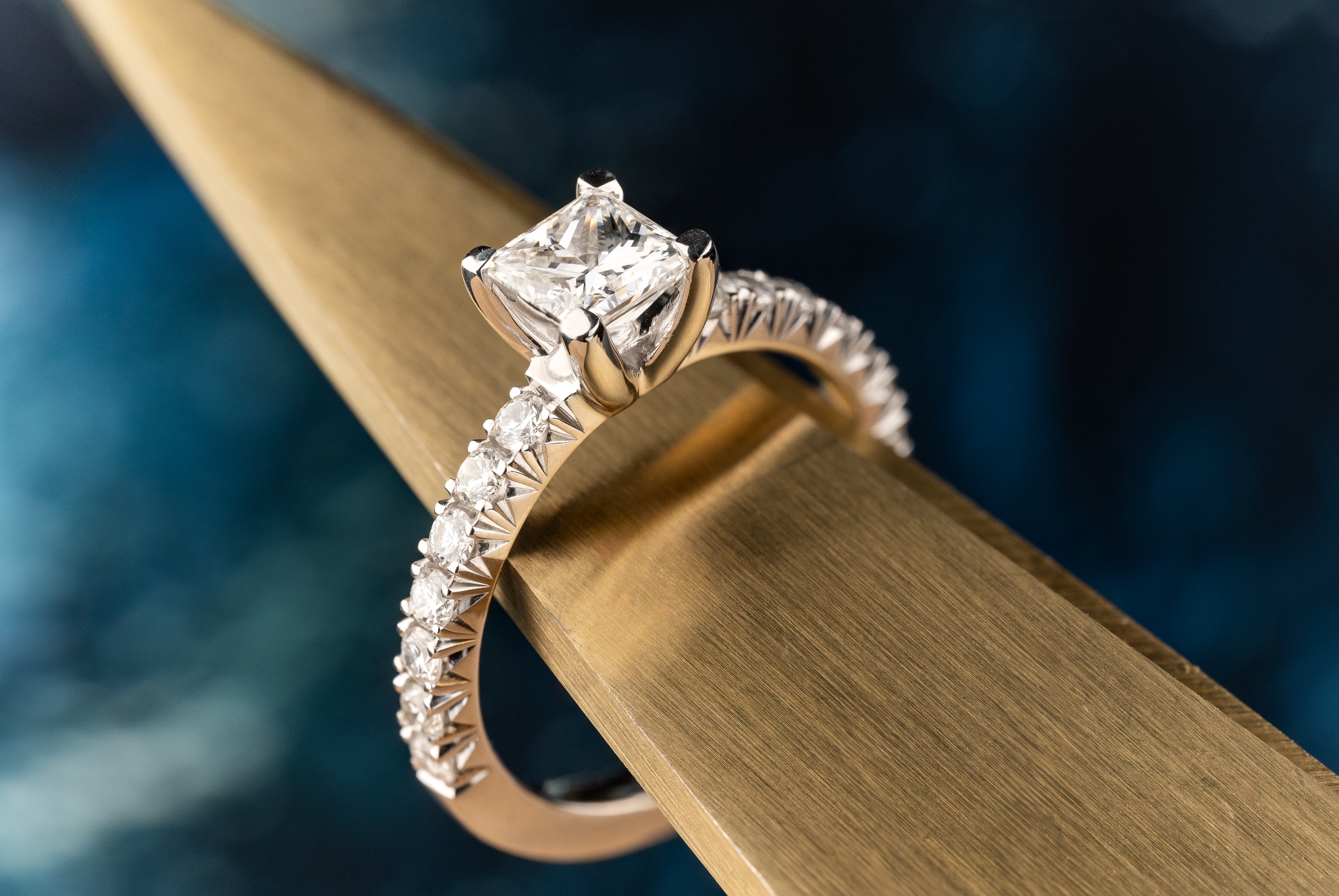 Pierścionek zaręczynowy z brylantem o szlifie princess, osadzony na złotej szynie z drobnymi diamentami, prezentowany na złotej, szczotkowanej powierzchni na tle ciemnoniebieskiego tła.