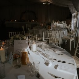 Elegancko nakryty stół weselny z białym obrusem, świecami, kwiatami i menu. W tle widoczne inne stoły i dekoracje, tworzące uroczystą atmosferę.