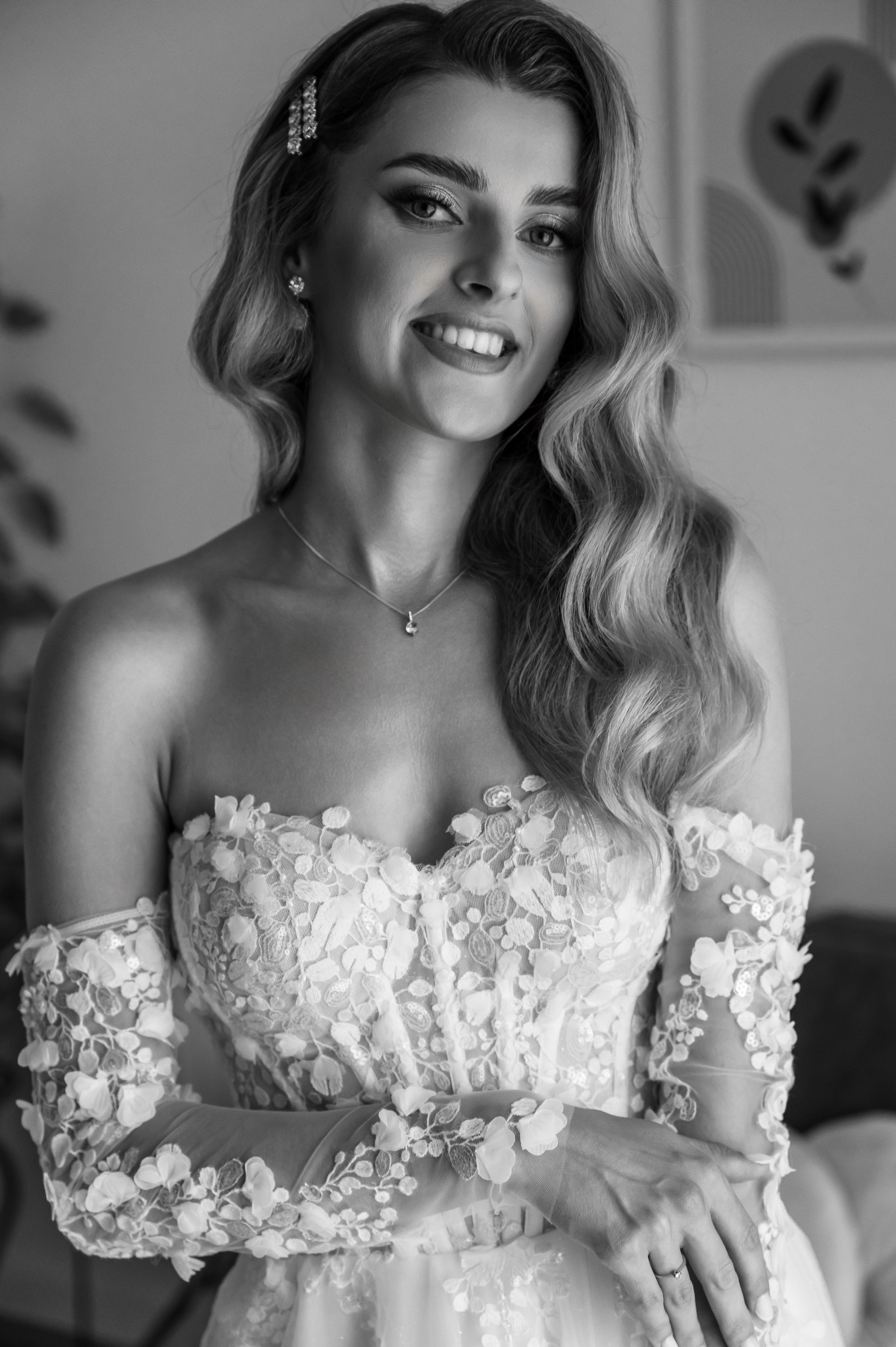 Uśmiechnięta panna młoda w koronkowej sukni z odkrytymi ramionami i ozdobą we włosach, czarno-białe zdjęcie.