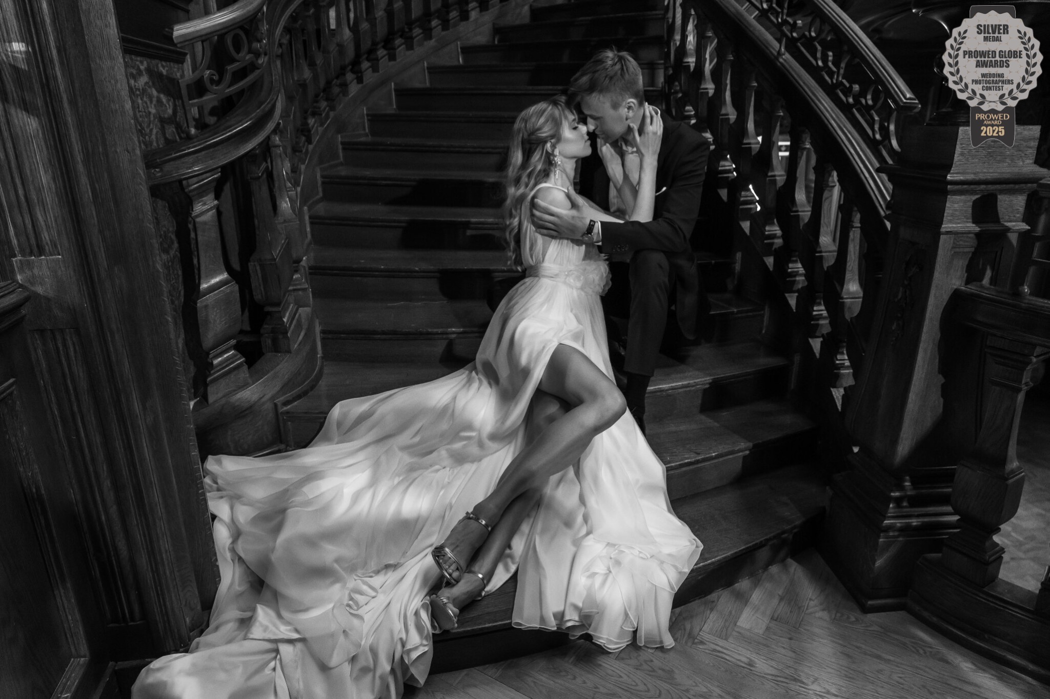 Czarno-białe zdjęcie pary młodej na drewnianych schodach z ozdobną balustradą. Panna młoda w sukni z rozcięciem, pan młody w garniturze. Zdjęcie nagrodzone w konkursie fotograficznym.