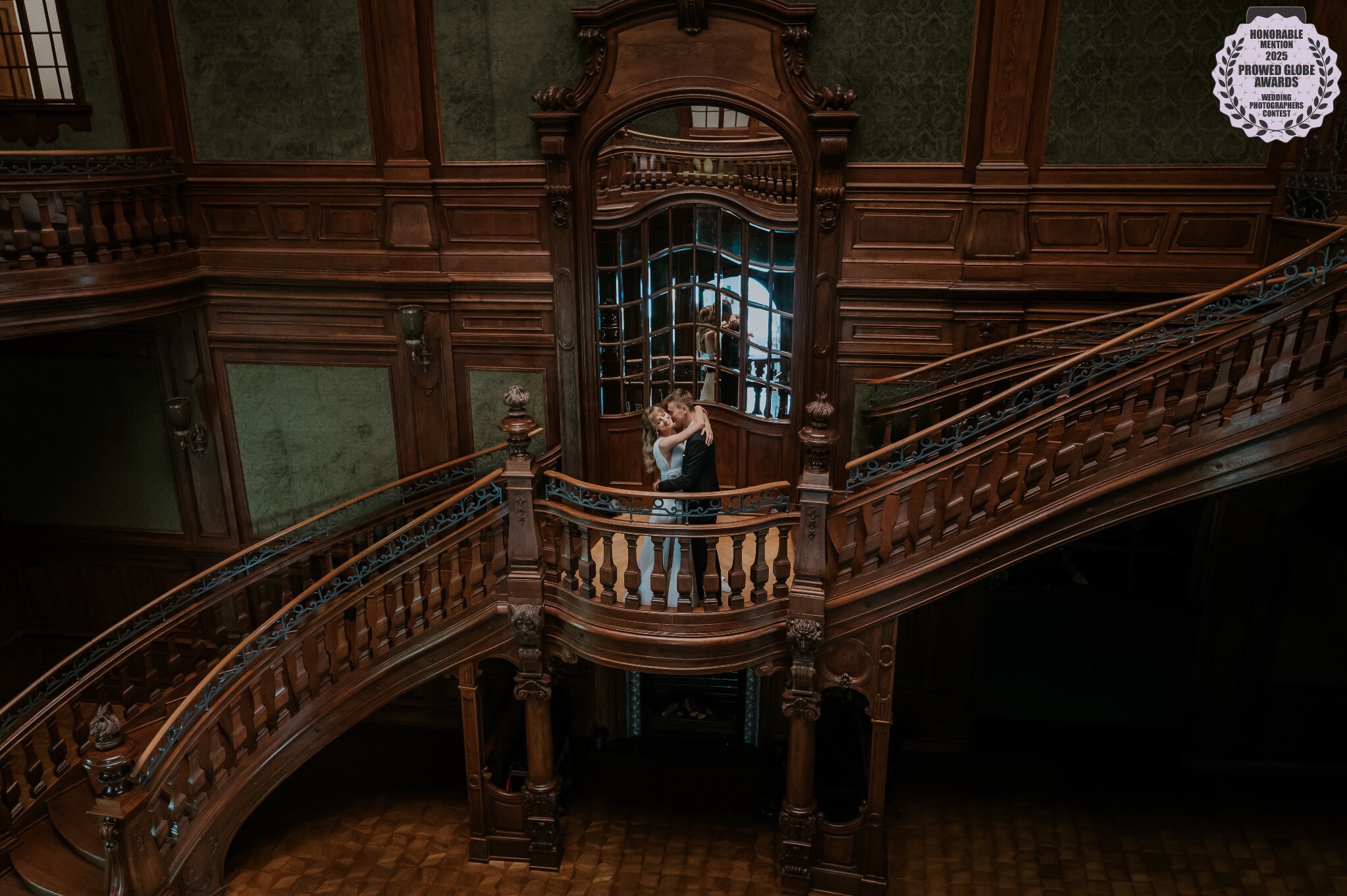 Para młoda w objęciach na tle bogato zdobionych, drewnianych schodów w zabytkowym wnętrzu. Widok z góry, nagroda fotograficzna w rogu.