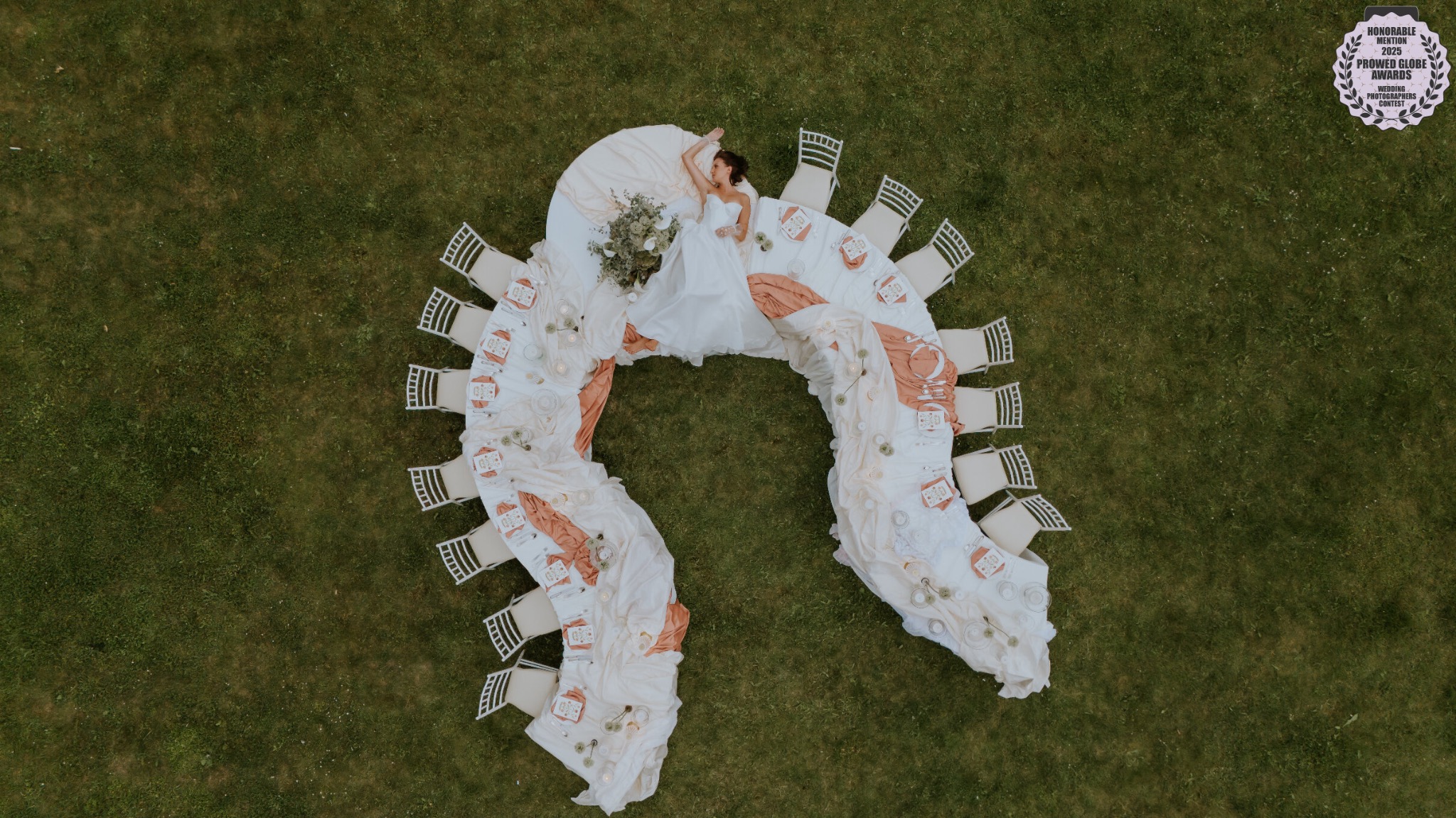 Kreatywne ujęcie z drona: Panna młoda w sukni leży na stole w kształcie podkowy, otoczonym krzesłami na zielonym trawniku. Dekoracje stołu w odcieniach bieli i różu.