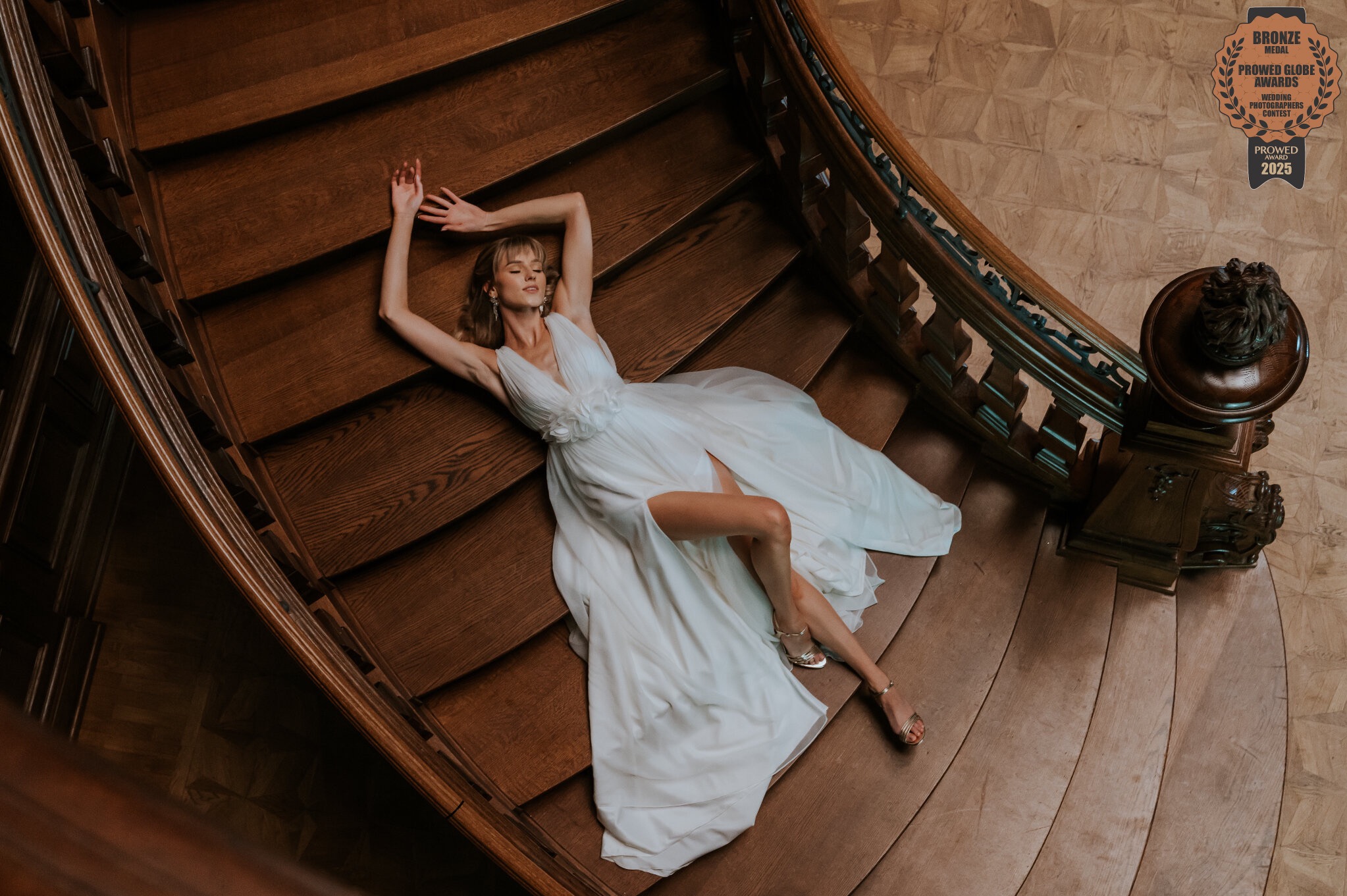 Kobieta w białej sukni leży na drewnianych schodach. Ujęcie z góry, widoczny fragment balustrady i podłogi. Na zdjęciu widnieje logo nagrody fotograficznej.
