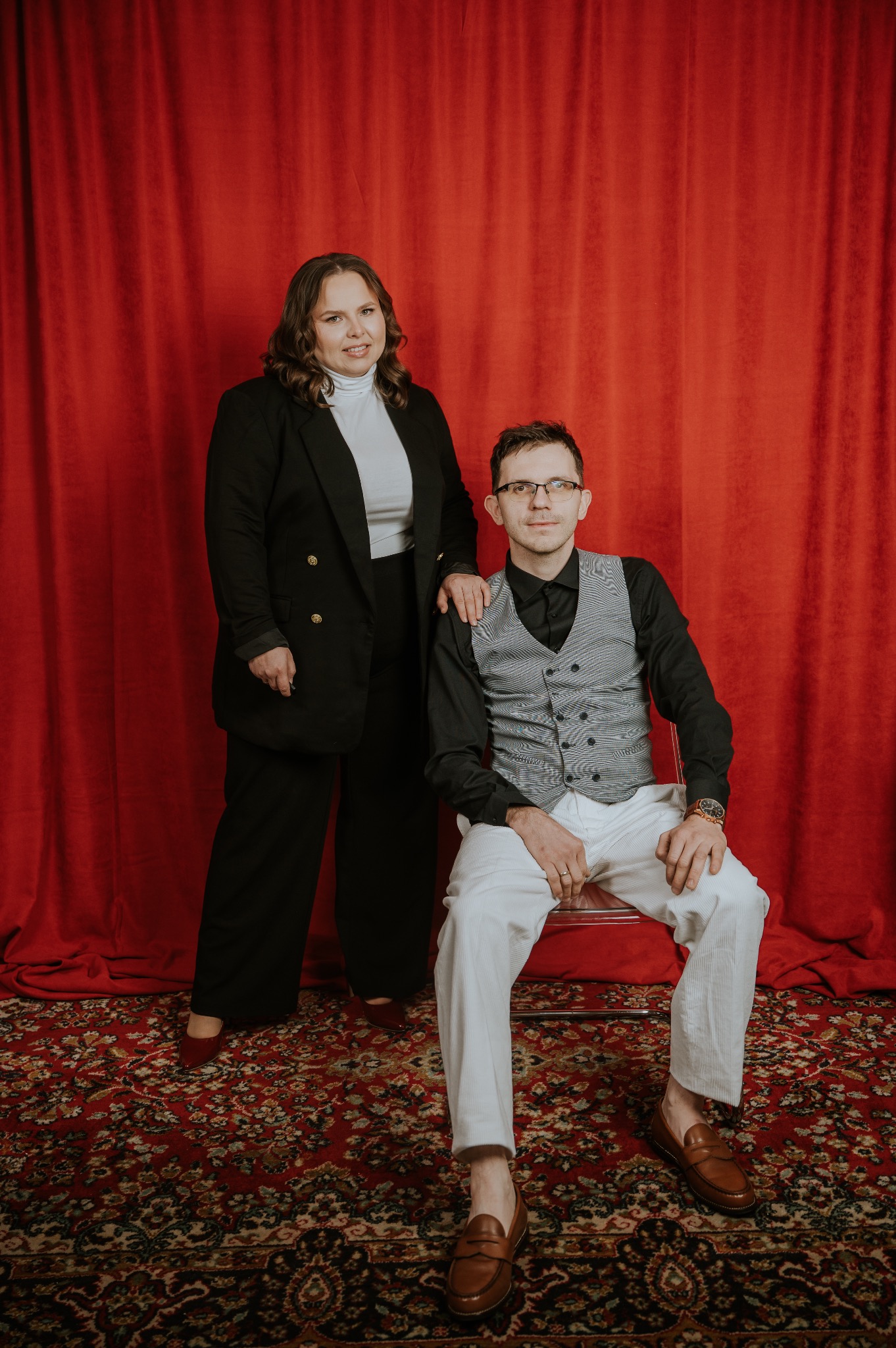 Portret biznesowy: kobieta w czarnym żakiecie stoi za siedzącym mężczyzną w kamizelce na tle czerwonej draperii i wzorzystego dywanu. Ujęcie studyjne, ciepłe światło.