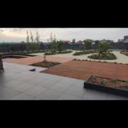 Nowoczesny taras z szarych płyt połączony z drewnianymi podestami i rabatami, w tle ogród z młodymi drzewkami i ciemnym ogrodzeniem.