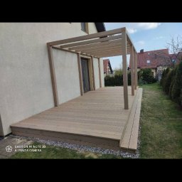 Jakub Bartosiak - Drewniany taras przylegający do domu z pergolą o jasnobeżowym odcieniu, widok z boku, z widoczną trawą i krzewami w tle.