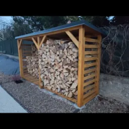 Drewniana wiata na drewno kominkowe z dachem, wypełniona pociętymi polanami, stojąca na żwirowym podłożu przy ogrodzeniu.