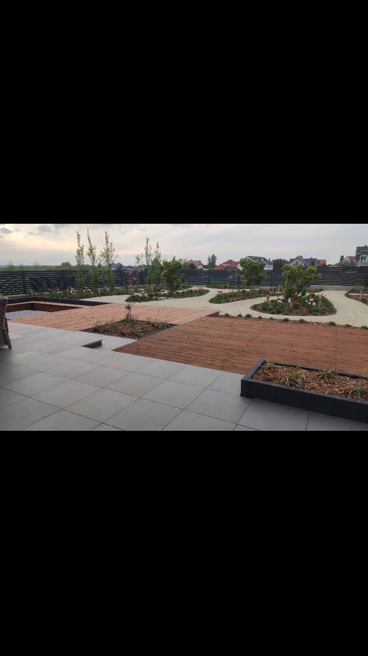 Nowoczesny taras z szarych płyt połączony z drewnianymi podestami i rabatami, w tle ogród z młodymi drzewkami i ciemnym ogrodzeniem.