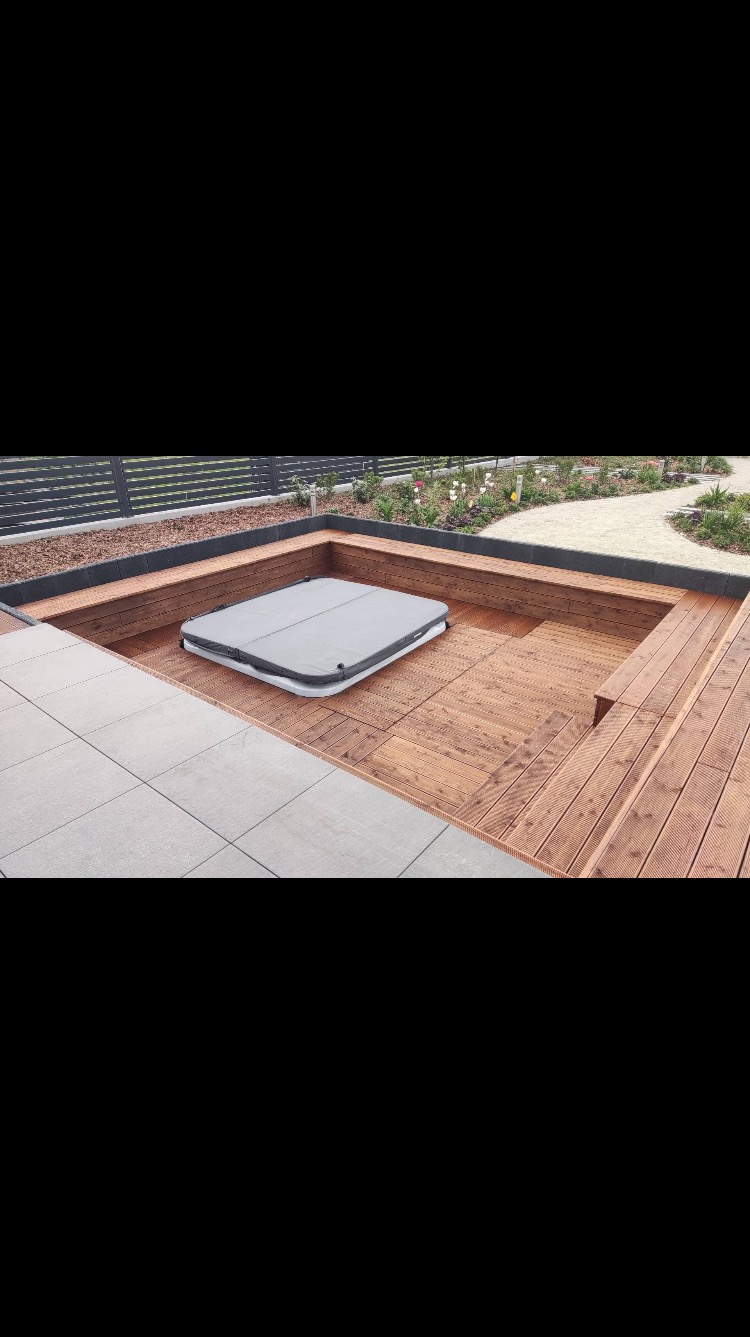 Zagłębiony taras z drewna z wbudowanym jacuzzi przykrytym szarą pokrywą, otoczony płytkami i rabatami kwiatowymi.