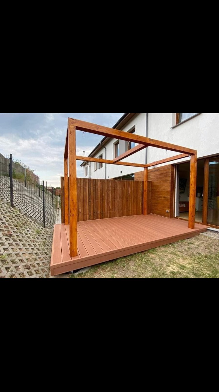 Nowoczesny taras kompozytowy z drewnianą konstrukcją zadaszenia, przylegający do budynku z drewnianą elewacją i dużymi oknami przesuwnymi, widok z boku na skarpę zabezpieczoną ażurowymi płytami...