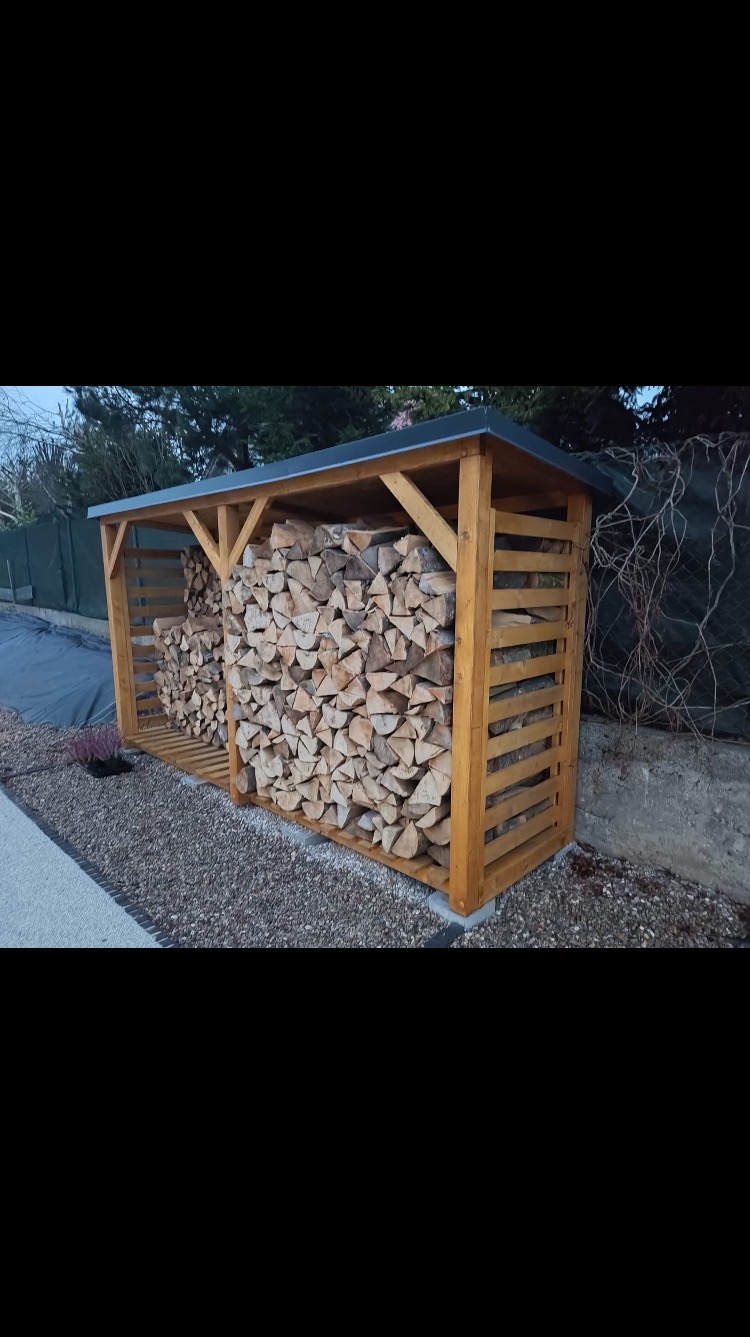 Drewniana wiata na drewno kominkowe z dachem, wypełniona pociętymi polanami, stojąca na żwirowym podłożu przy ogrodzeniu.