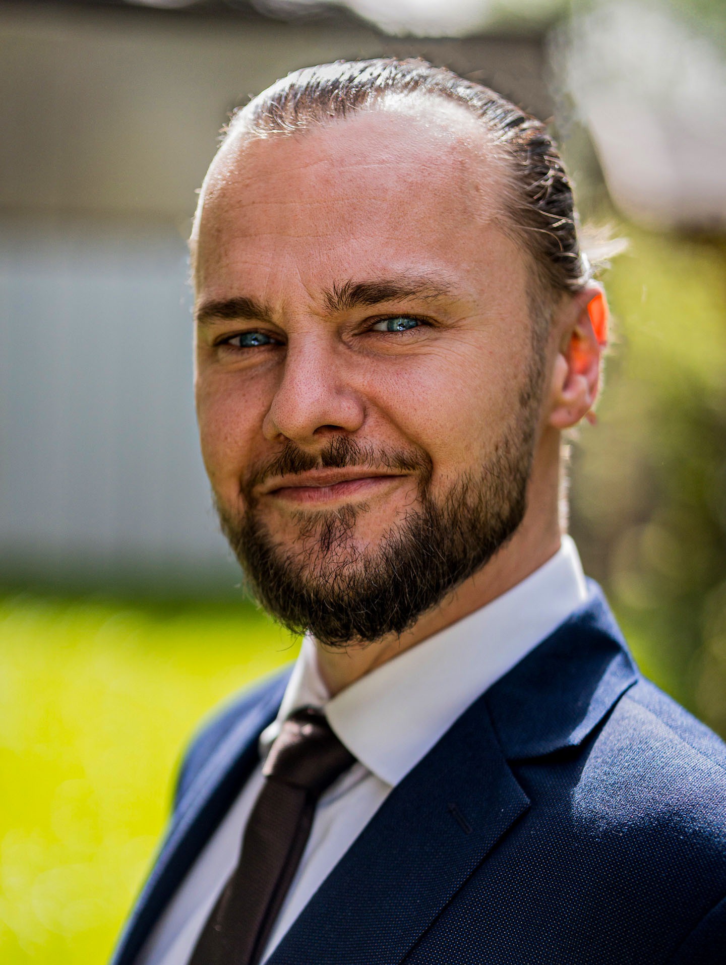 Portret mężczyzny w garniturze, z brodą i niebieskimi oczami, uśmiechającego się do kamery na tle zieleni w Poznaniu. Ujęcie z bliska, naturalne światło.