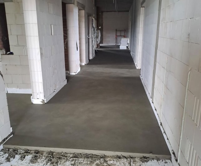 Wnętrze w trakcie wylewki posadzki jastrychowej, widoczne ściany z białego bloczka i świeżo wylana, gładka powierzchnia betonu.