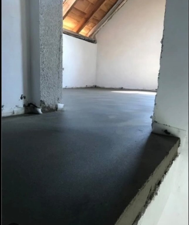 Świeżo wylana, gładka, szara posadzka w surowym stanie, z widocznym drewnianym stropem poddasza i fragmentami nieotynkowanych ścian.