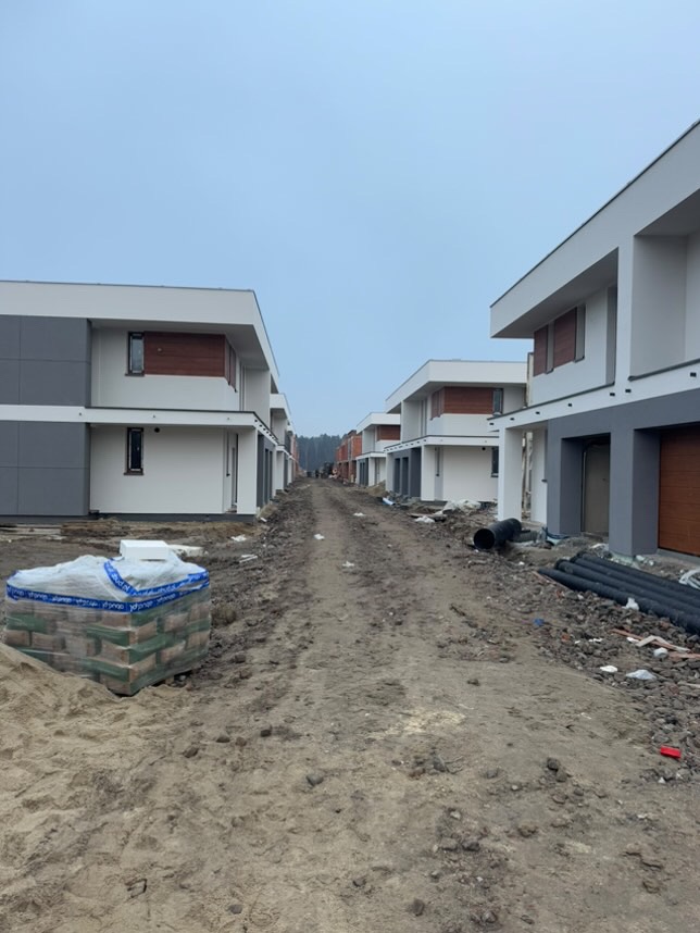 Ulica w trakcie budowy osiedla domów jednorodzinnych, widoczne materiały budowlane i rury na ziemi, domy w nowoczesnym stylu z elementami drewnianymi.