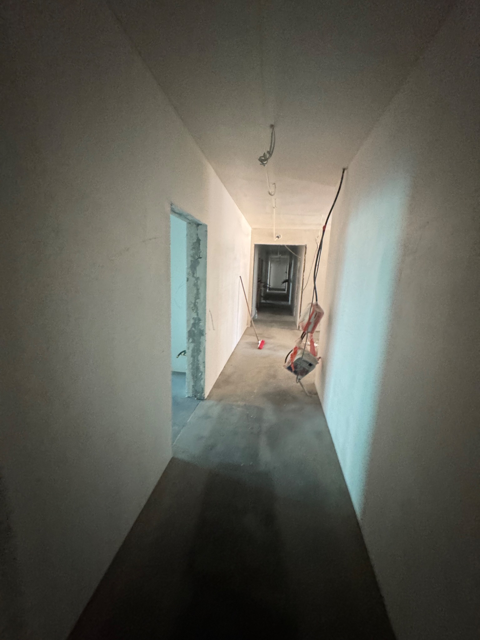 Długi korytarz w surowym stanie: szare ściany, betonowa posadzka, wiszące kable elektryczne. Widoczne niedokończone ościeżnice i narzędzia budowlane w oddali.