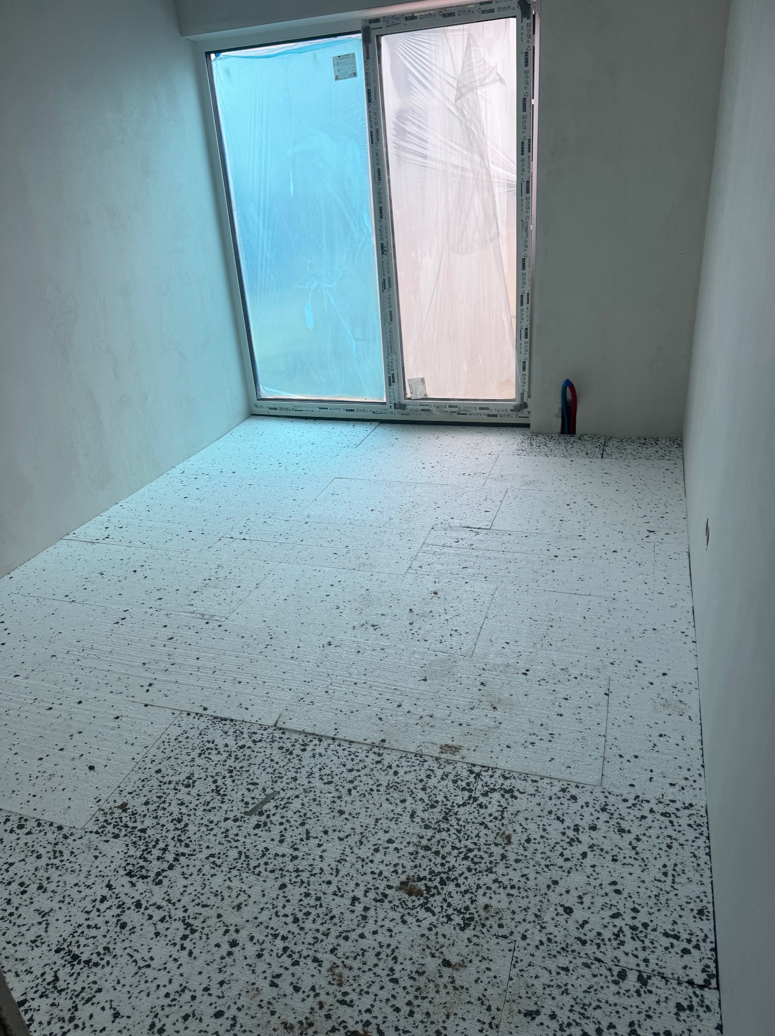 Przygotowanie podłoża pod posadzkę: rozłożone płyty izolacyjne na podłodze w pomieszczeniu z oknem zabezpieczonym folią. Widoczne ślady zabrudzeń.