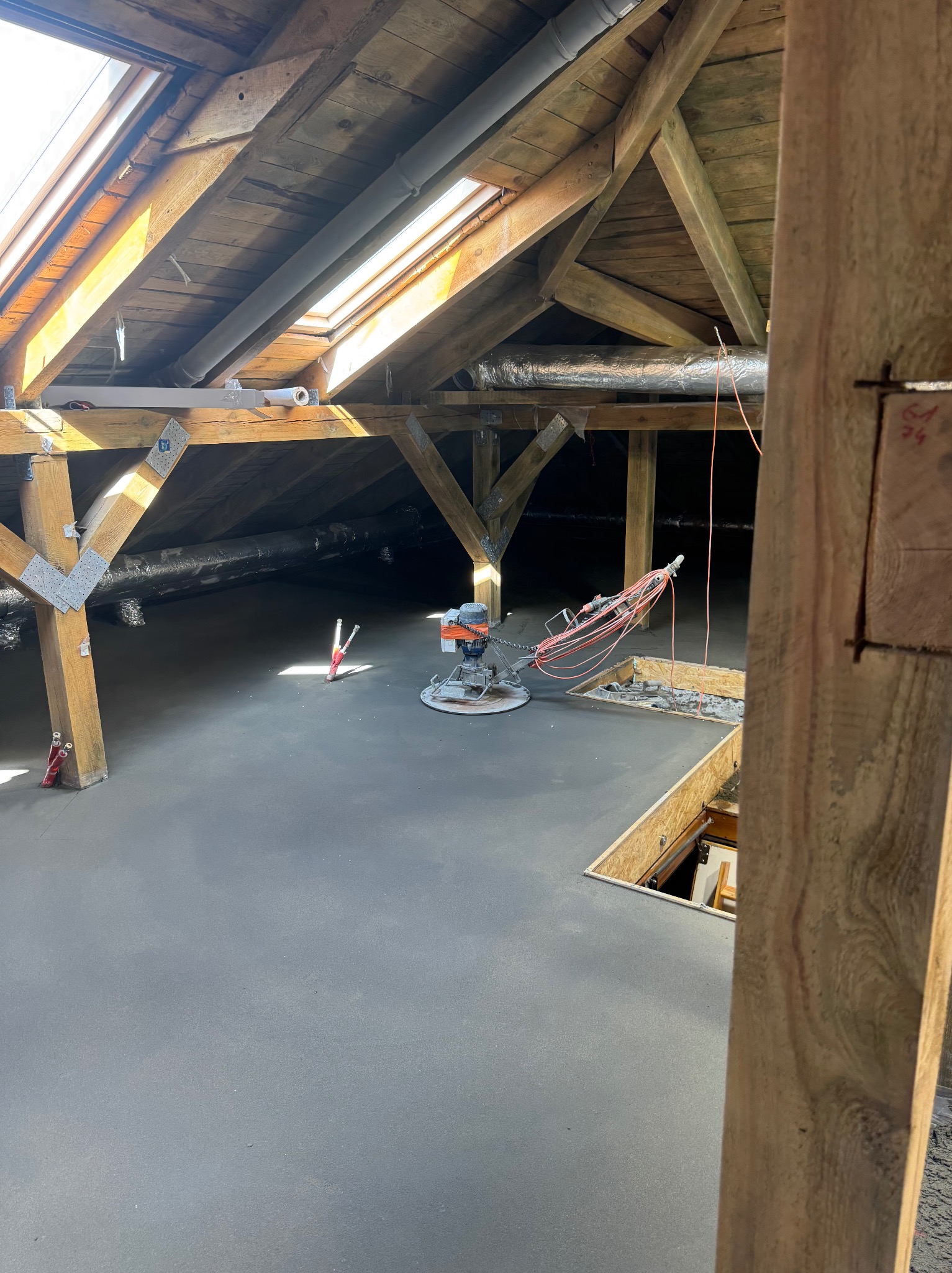 Szlifowanie posadzki jastrychowej na poddaszu z drewnianą konstrukcją. Widoczna maszyna do szlifowania i gładka, szara powierzchnia posadzki. Oświetlenie naturalne z okna dachowego.