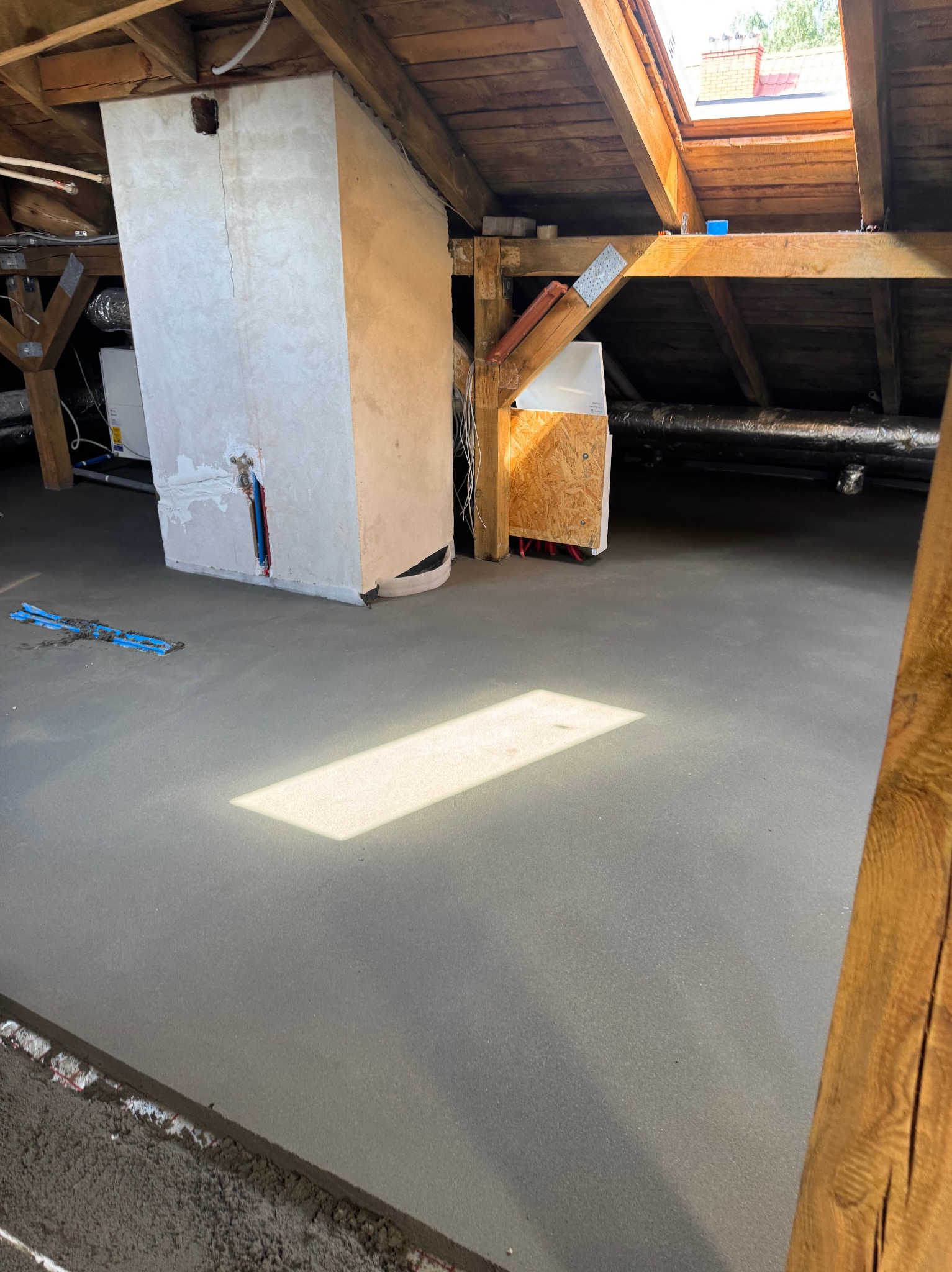 Świeżo wylana, gładka posadzka jastrychowa na poddaszu z drewnianą konstrukcją dachu i oknem dachowym. Widoczne elementy instalacji i fragment ściany.