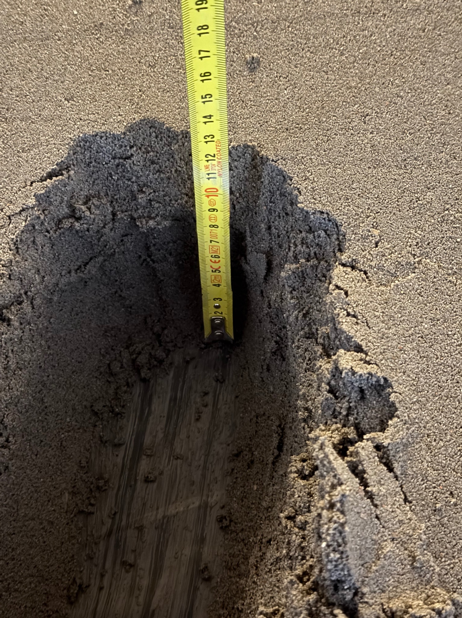 Pomiar głębokości wnęki w szarej, ziarnistej powierzchni za pomocą żółtej miarki. Widoczne ślady po narzędziu. Bliski kadr, ostre światło.